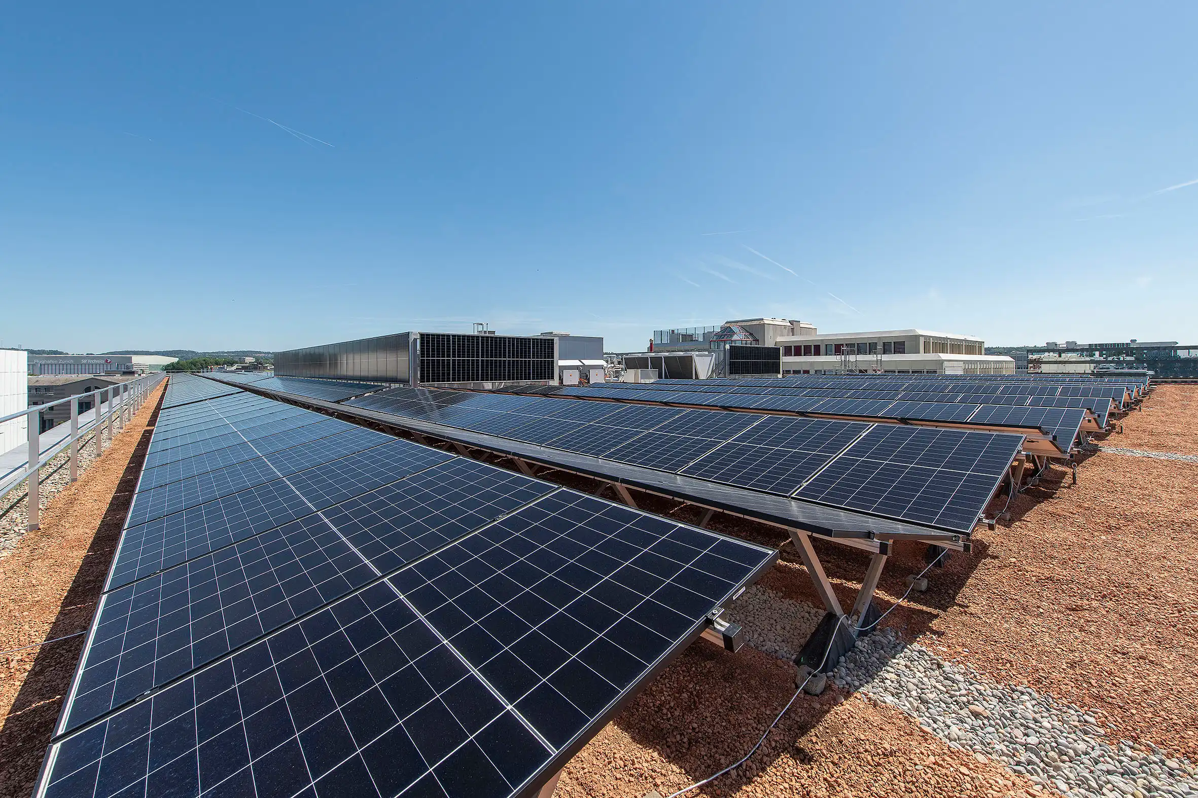 Solar panels arranged in rows on a gravel rooftop under a clear blue sky, with buildings in the distance.