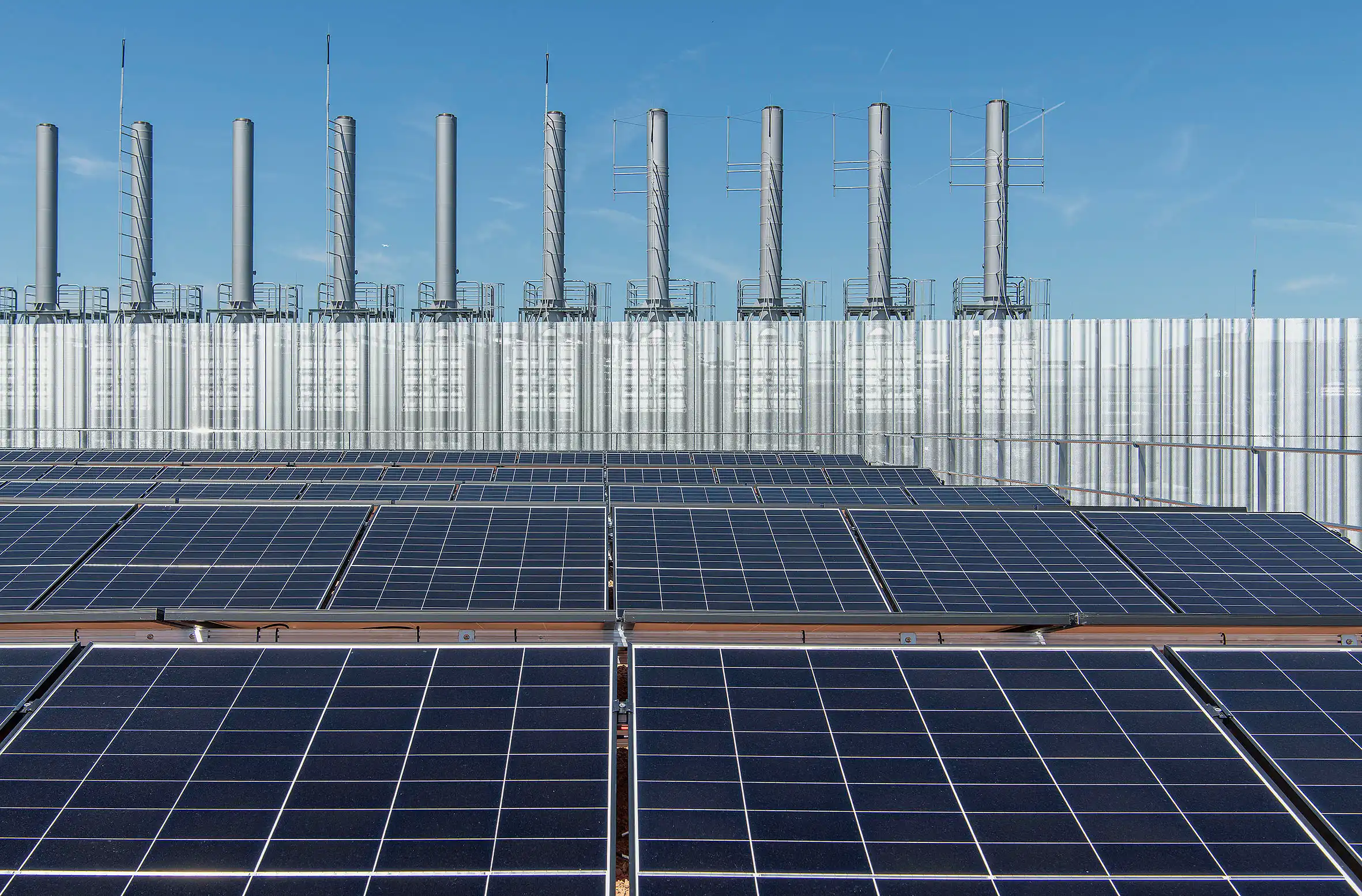 Row of dark solar panels on a rooftop with tall gray ventilation stacks against a clear blue sky.