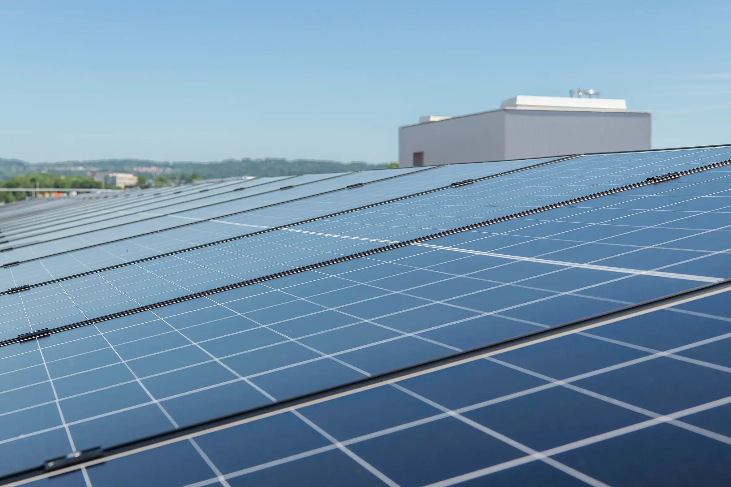 Rooftop solar panels arranged in neat rows under a clear blue sky with a small building in the background.