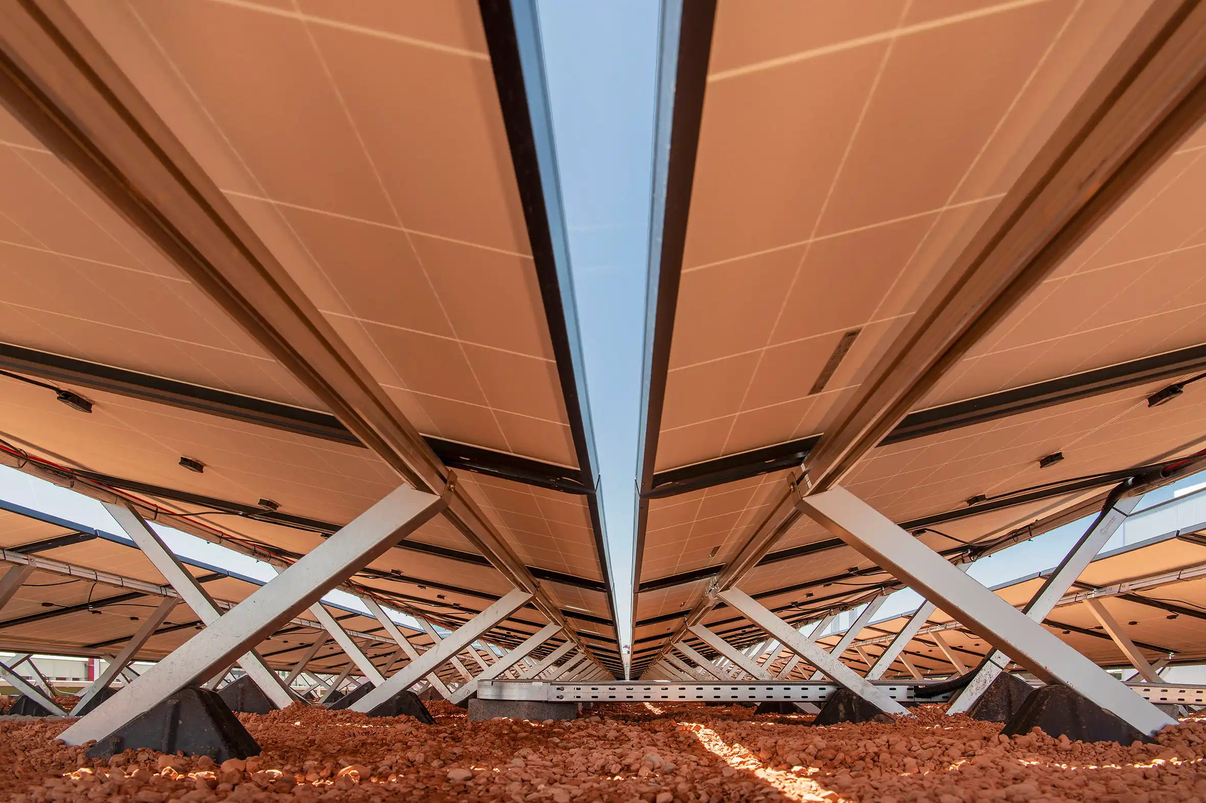 View upward at a construction site: orange panel roof supported by white steel trusses and dark beams, viewed from ground amid gravel. Perspective from below shows intersecting metal beams forming a tunnel-like structure with a sky gap at center.