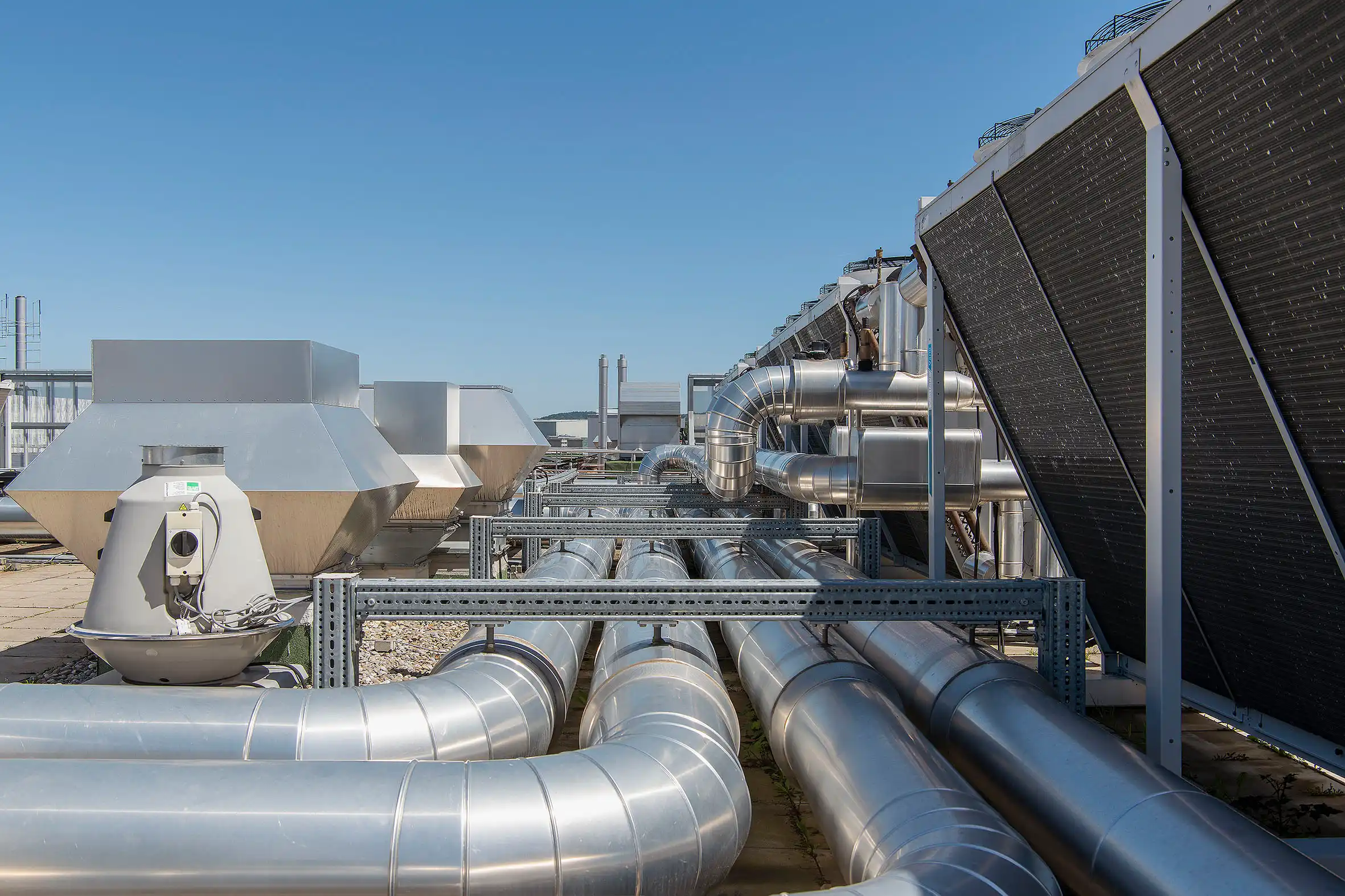 Industrial rooftop with interconnected silver HVAC ducts and vents, beside dark solar panels against a blue sky.