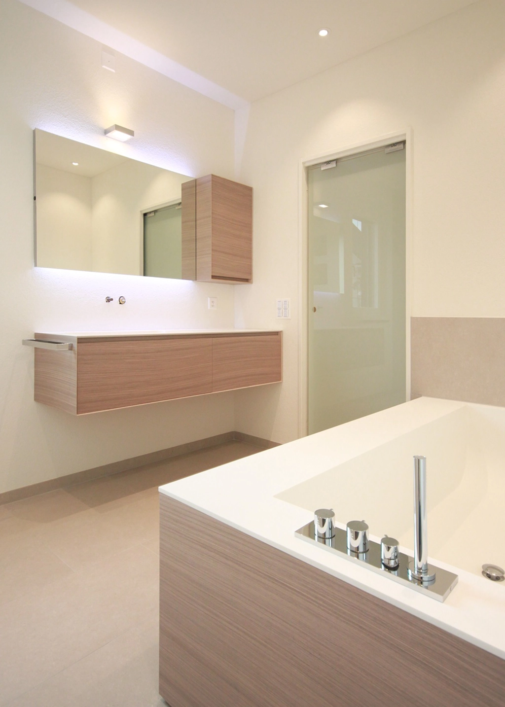 Modern bathroom with a floating wooden vanity, wall cabinet, large mirror, and frosted glass door next to a white tub with chrome faucets.
