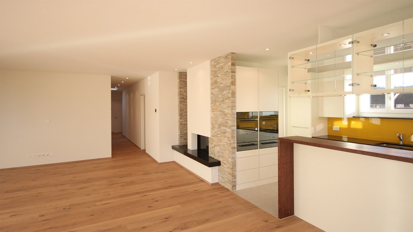 Open-concept living space with wood floors, white walls, and a modern kitchen featuring a stone-clad column and built-in fireplace alcove on the left.