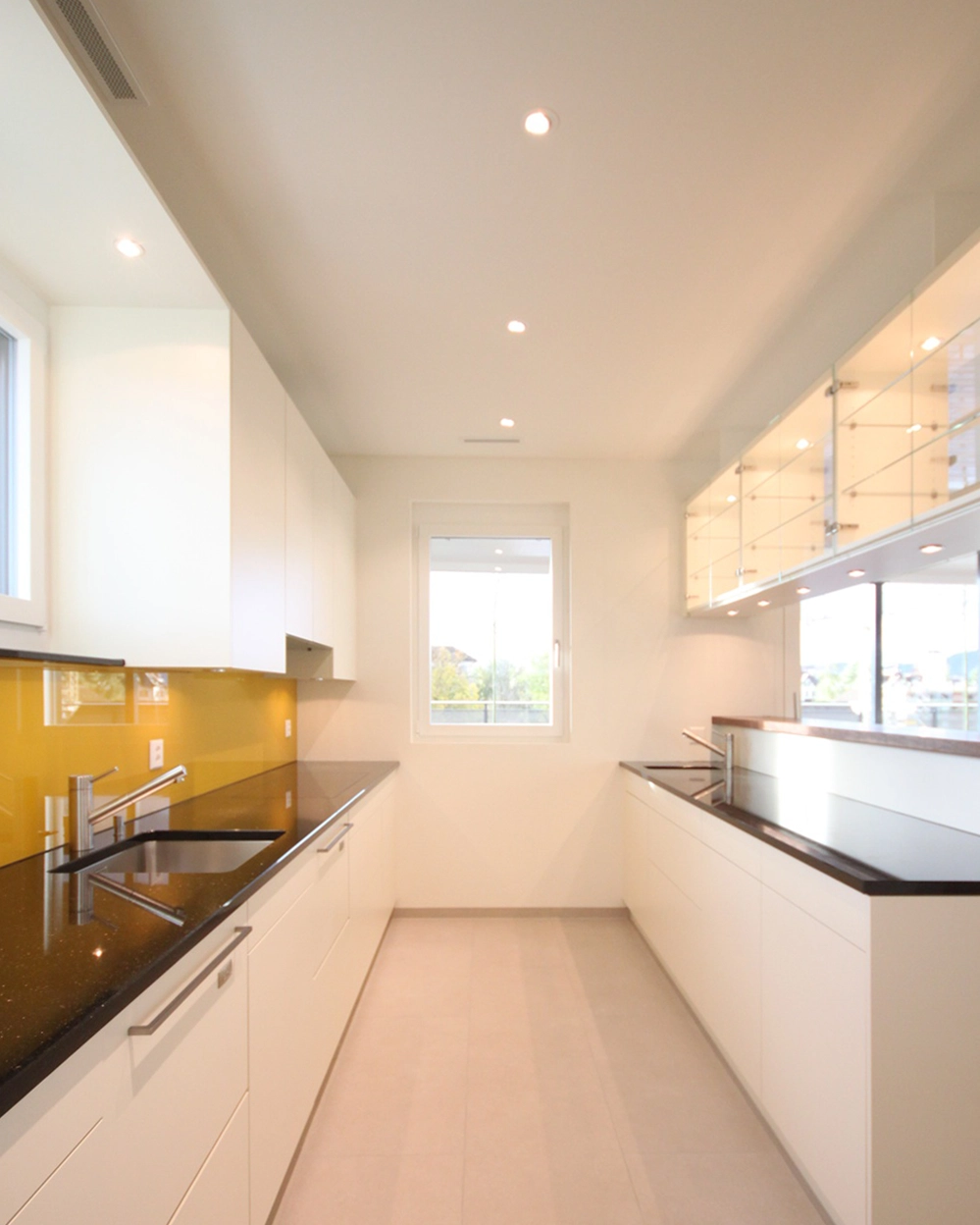 Bright modern galley kitchen with white cabinets, black countertops, yellow backsplash, and a window at the far end.