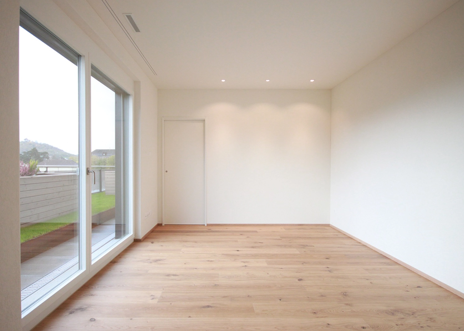Bright empty living room with light wood floor, white walls, and large glass doors opening to a balcony outside.
