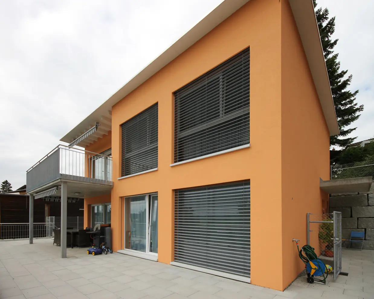 Orange two-story house with large, horizontal window blinds, a balcony on the left, and a tiled patio with outdoor furniture and toys.