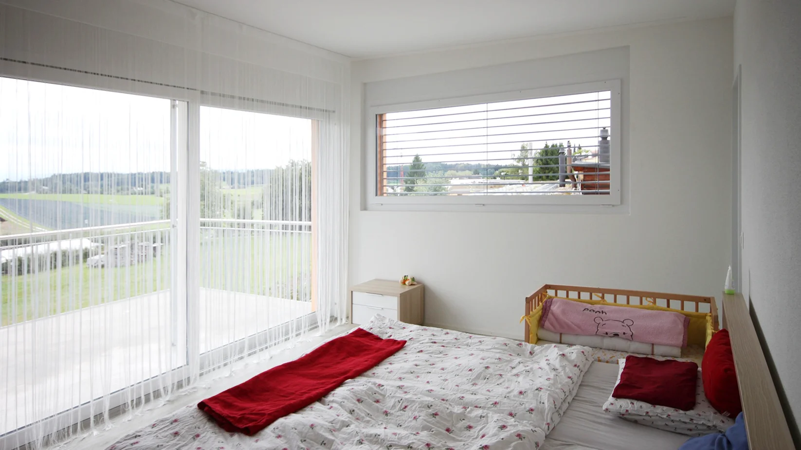 Bright bedroom with a low wooden bed, floral bedding, red throw, and a pink baby cot along the right wall; large sliding doors with sheer blinds.