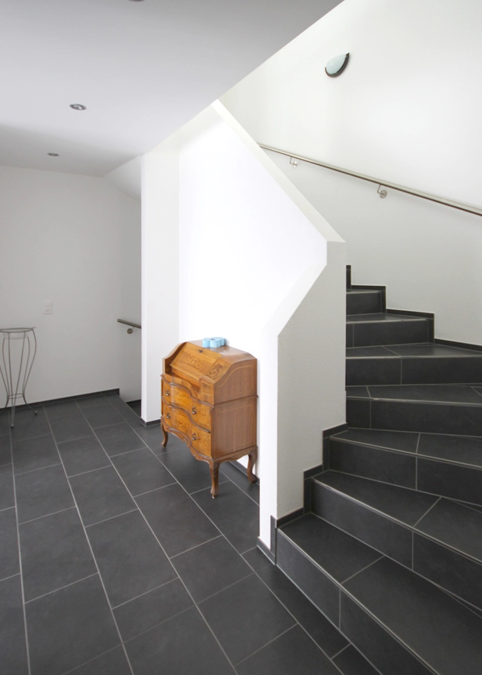 Interior entry area with a white staircase, dark floor tiles, and a wooden antique dresser beside a white pillar.