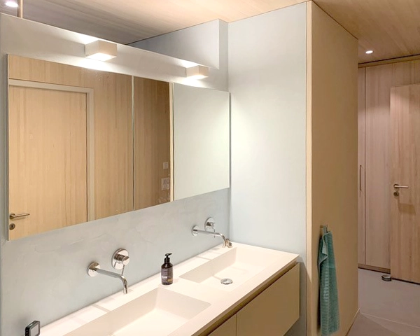 Double-sink bathroom vanity with wall-mounted faucets, a large mirror, soap dispenser, and a turquoise towel by a light wood door.