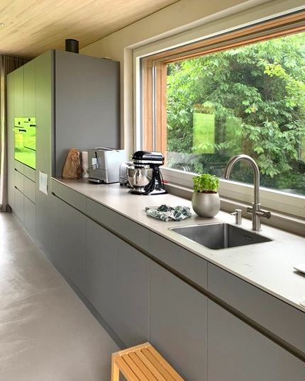 Modern grey kitchen with a long counter, large window showing greenery, and a sink with a plant nearby.