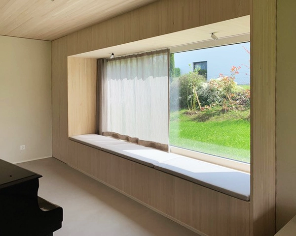 Room with a built-in window seat along a large bay window, light wood paneling, and sheer curtains; garden visible outside.