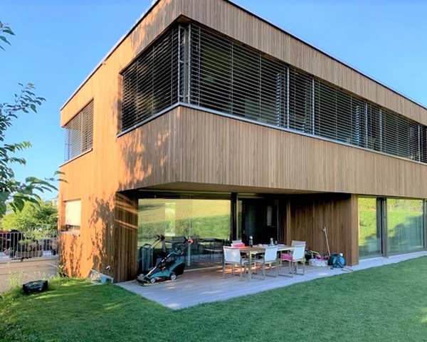 Modern two-story wooden house with large glass doors; outdoor dining table and chairs on a concrete patio, green lawn.