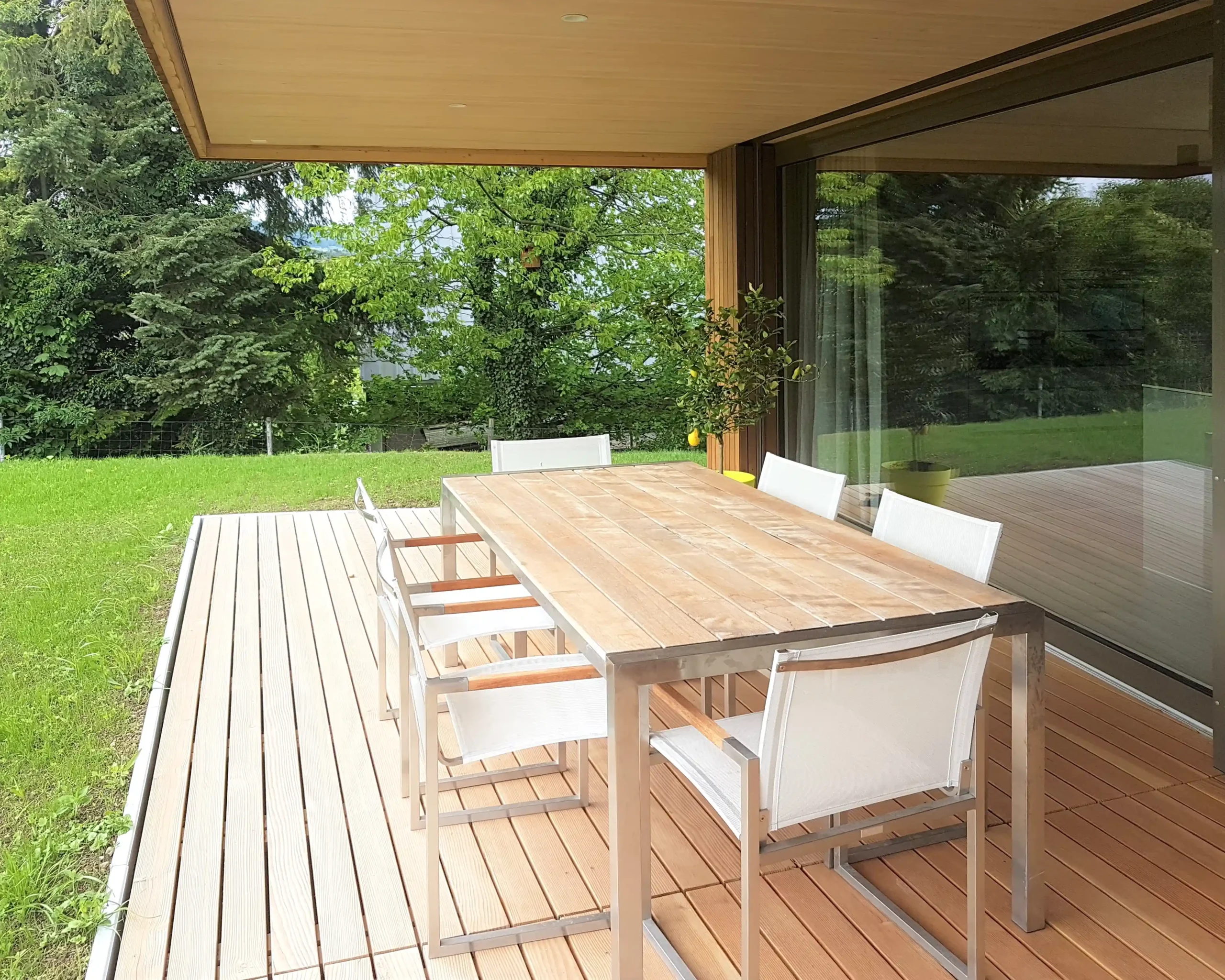 Outdoor wooden deck with a long table and white chairs under a covered patio, green lawn beyond.