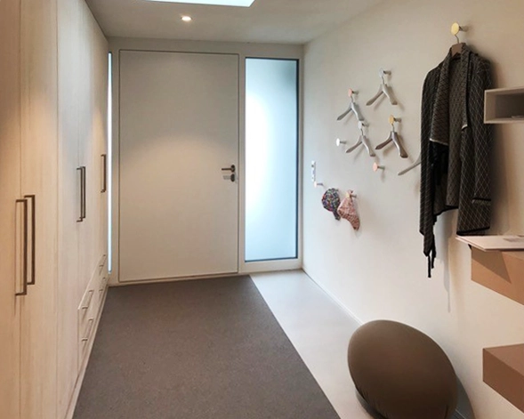 Narrow modern entryway with built-in wardrobes on the left, white walls, and coat hooks on the right; a dark robe hangs and a rounded brown pouf sits near the wall, with a gray runner rug leading to a frosted glass door at the end.
