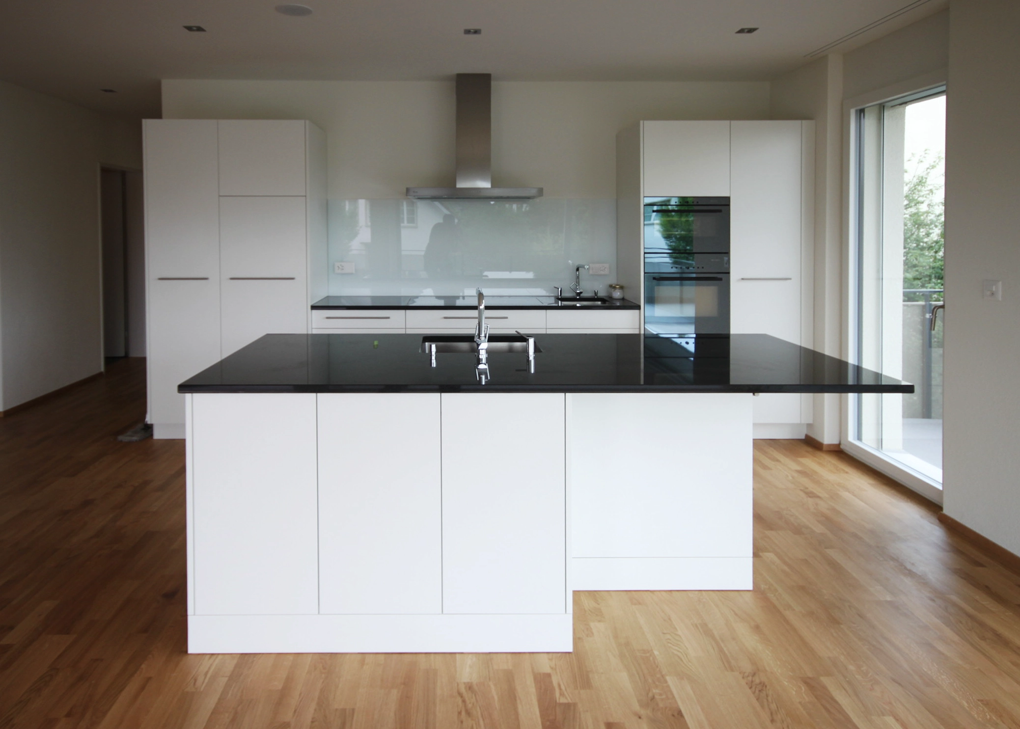 Modern white kitchen with a central black countertop island, stainless steel range hood, built-in double ovens, and white cabinetry.