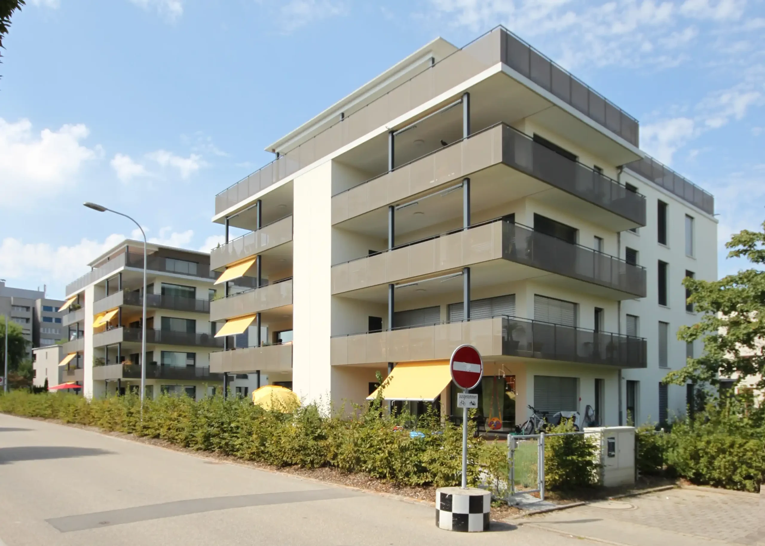 Modern multi-story apartment building with glass balconies along a sunny street.