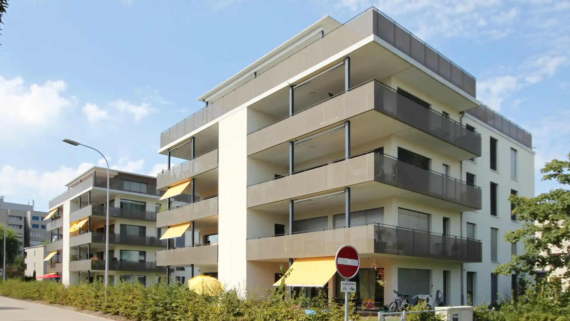 Modern white apartment building with stacked balconies and glass railings under a blue sky. A street lamp and yellow awnings are visible on the ground floor.