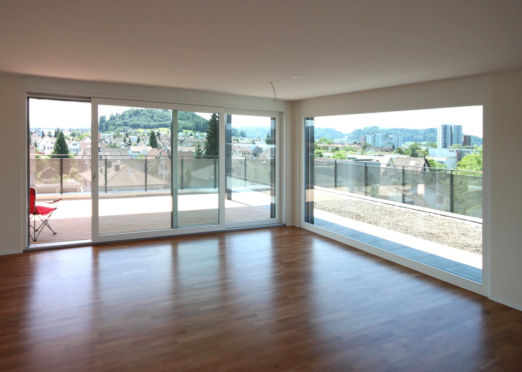 Bright empty living room with wide sliding glass doors opening to a balcony and a town view, hills beyond. (informative)