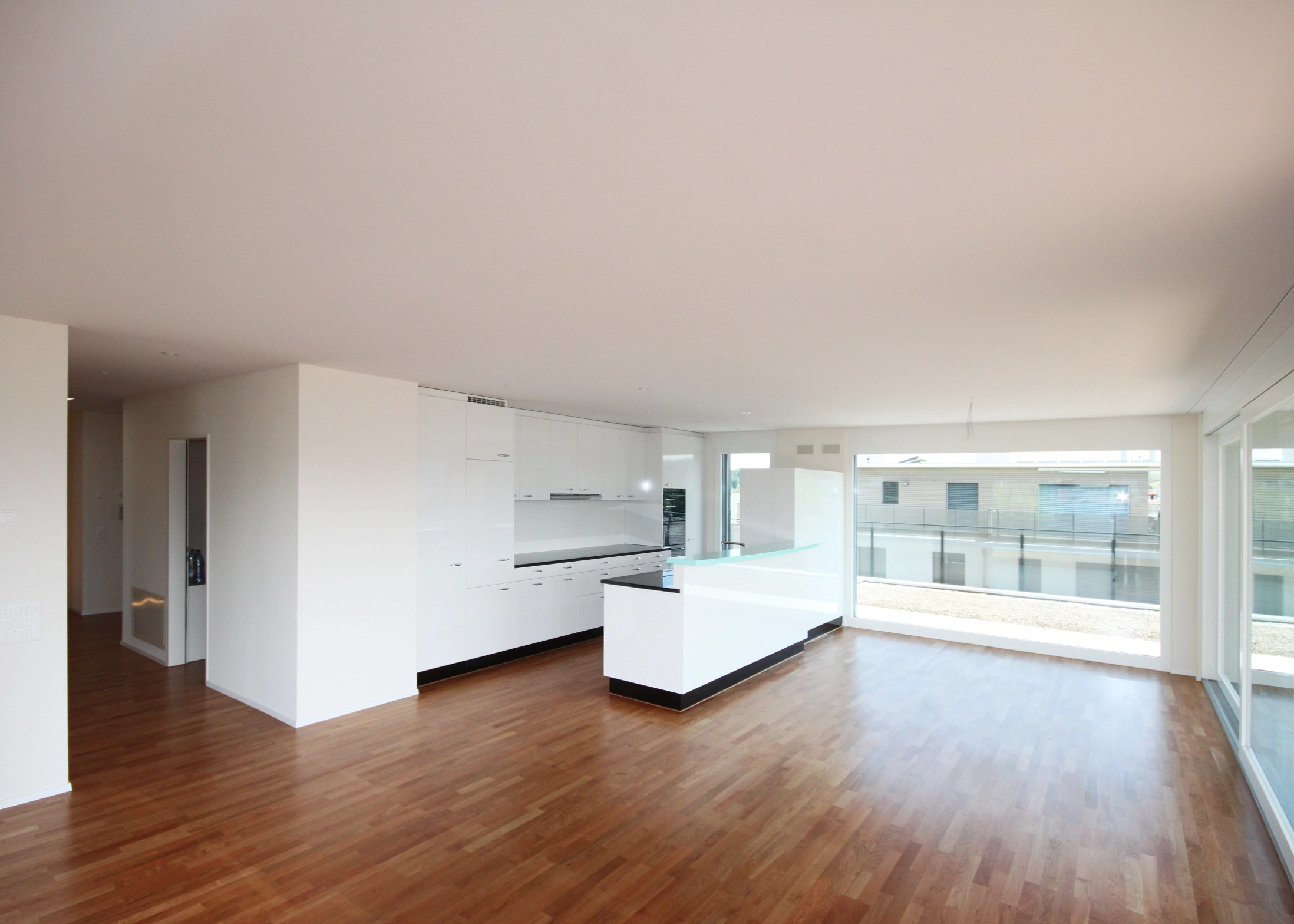 Bright open-plan kitchen with white cabinetry and glass accents, large windows overlooking buildings, and warm wooden flooring.