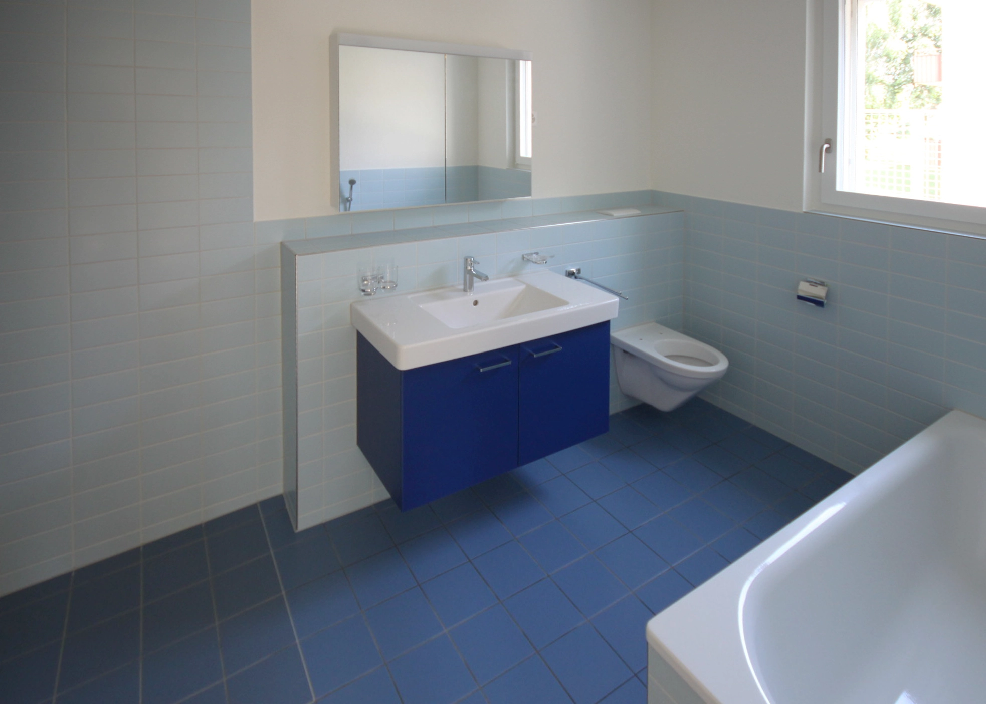 Blue-tiled bathroom with a white rectangular sink on a blue cabinet, mirrored wall cabinet, toilet, and a window above the tub rim.