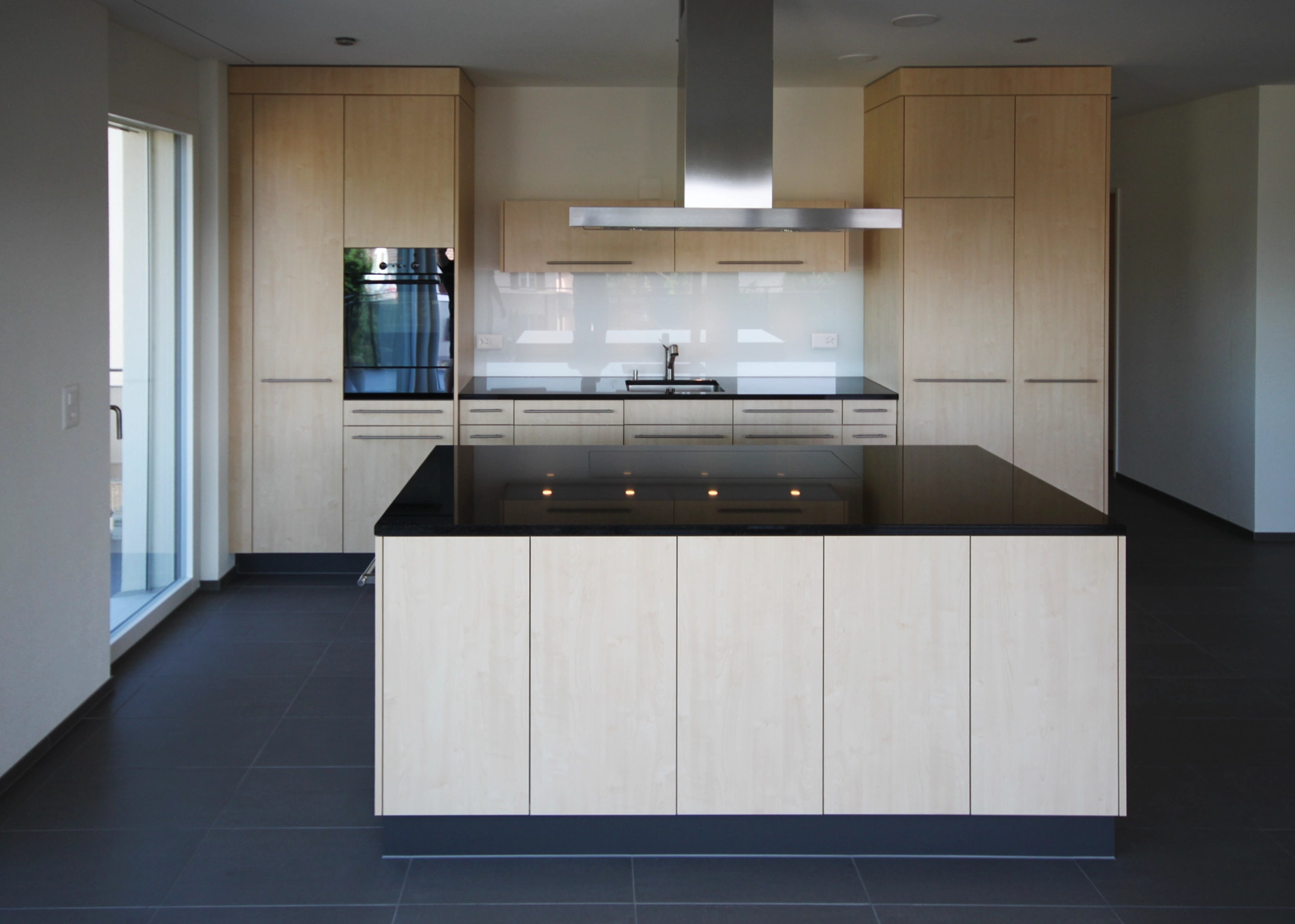 Modern kitchen with light wood cabinets, black countertops, and a central island with a stainless range hood above the stove.