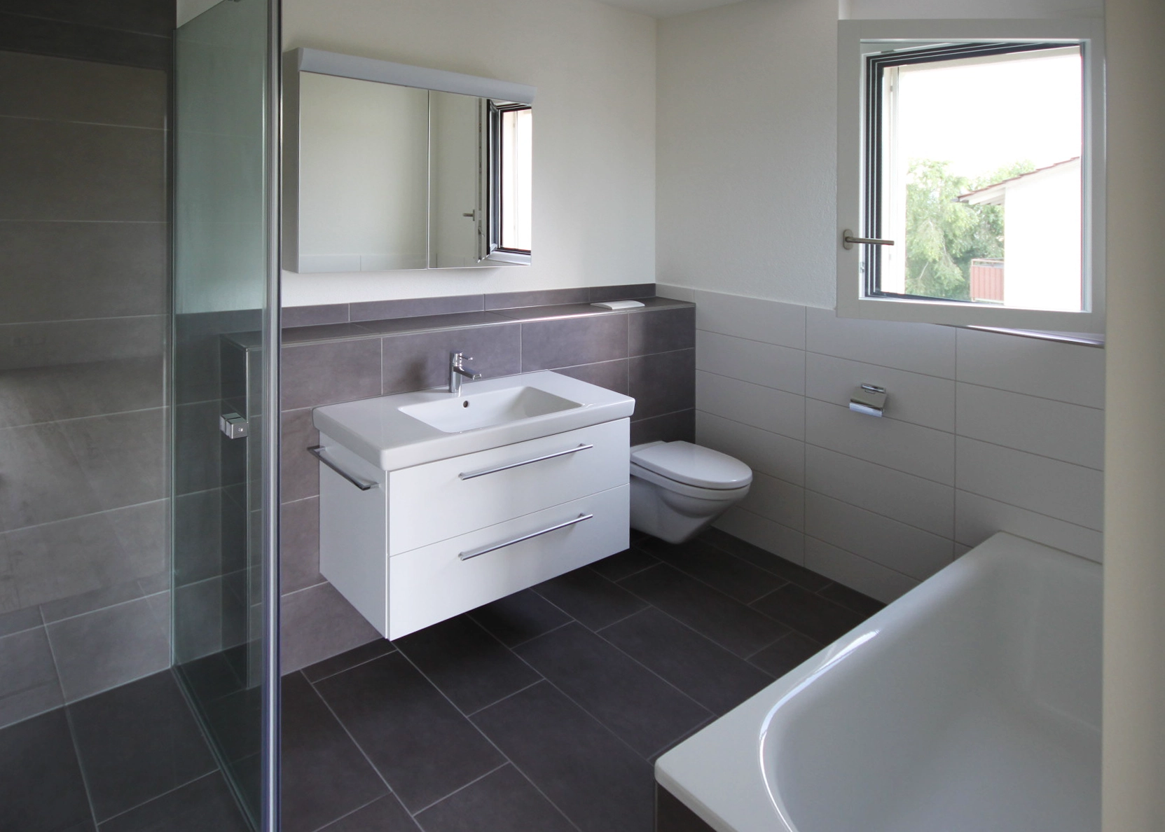 Modern bathroom with white vanity, rectangular sink, and wall mirror; gray tiled walls and floor, glass shower on left, toilet beside vanity, and a window for natural light.