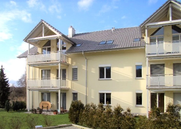Three-story beige apartment building with balconies and a gabled roof, set in a manicured lawn and shrubs.