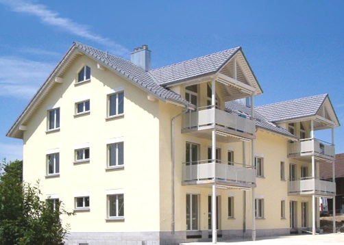 Three-story yellow apartment building with white balconies and a tiled roof under a blue sky.