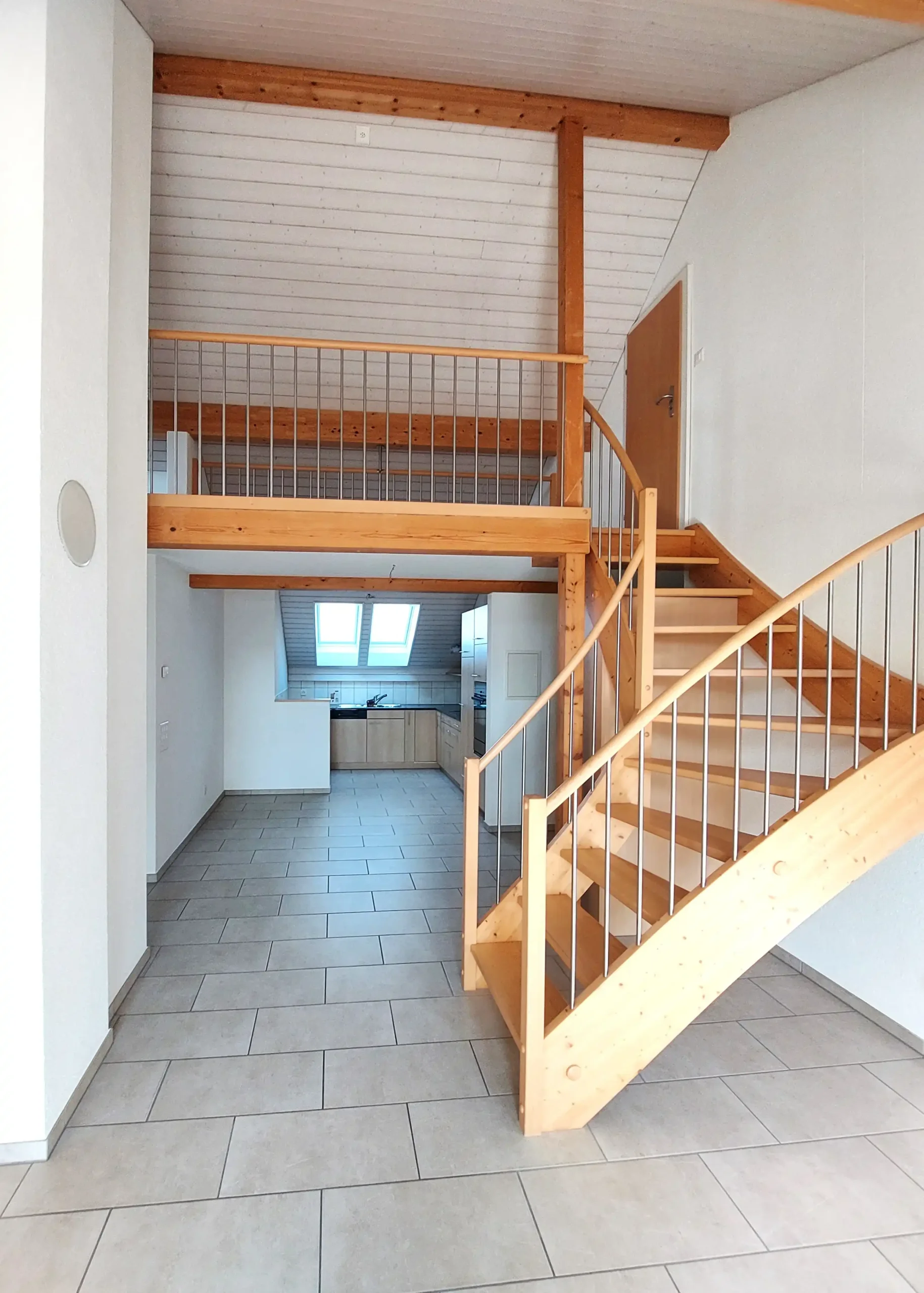 Open-plan interior with a wooden staircase and loft railing; kitchen visible in the background through a doorway.