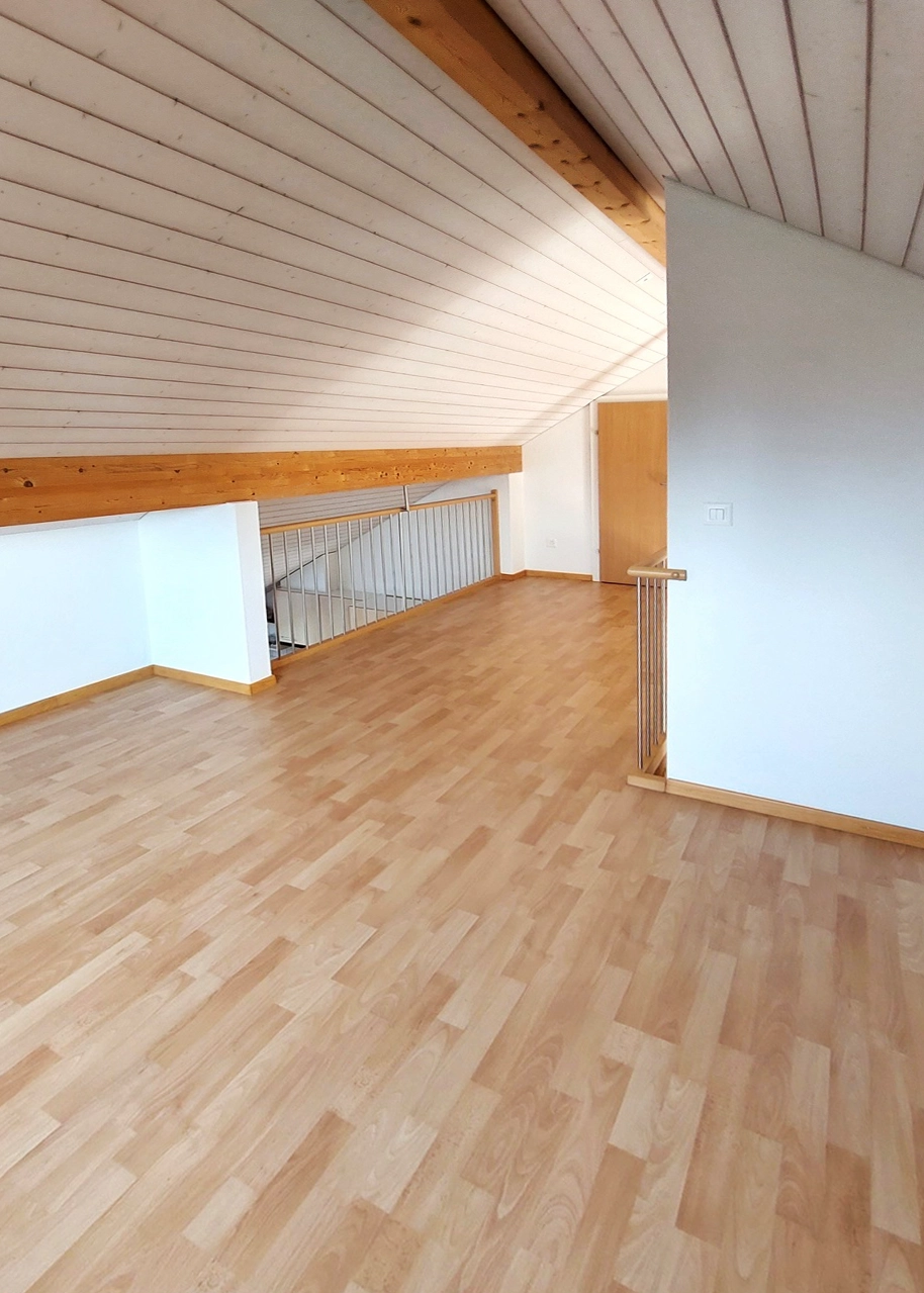 Empty attic loft with a light wood floor, white walls, and a slanted wooden ceiling with exposed beams; railing overlooks a staircase.