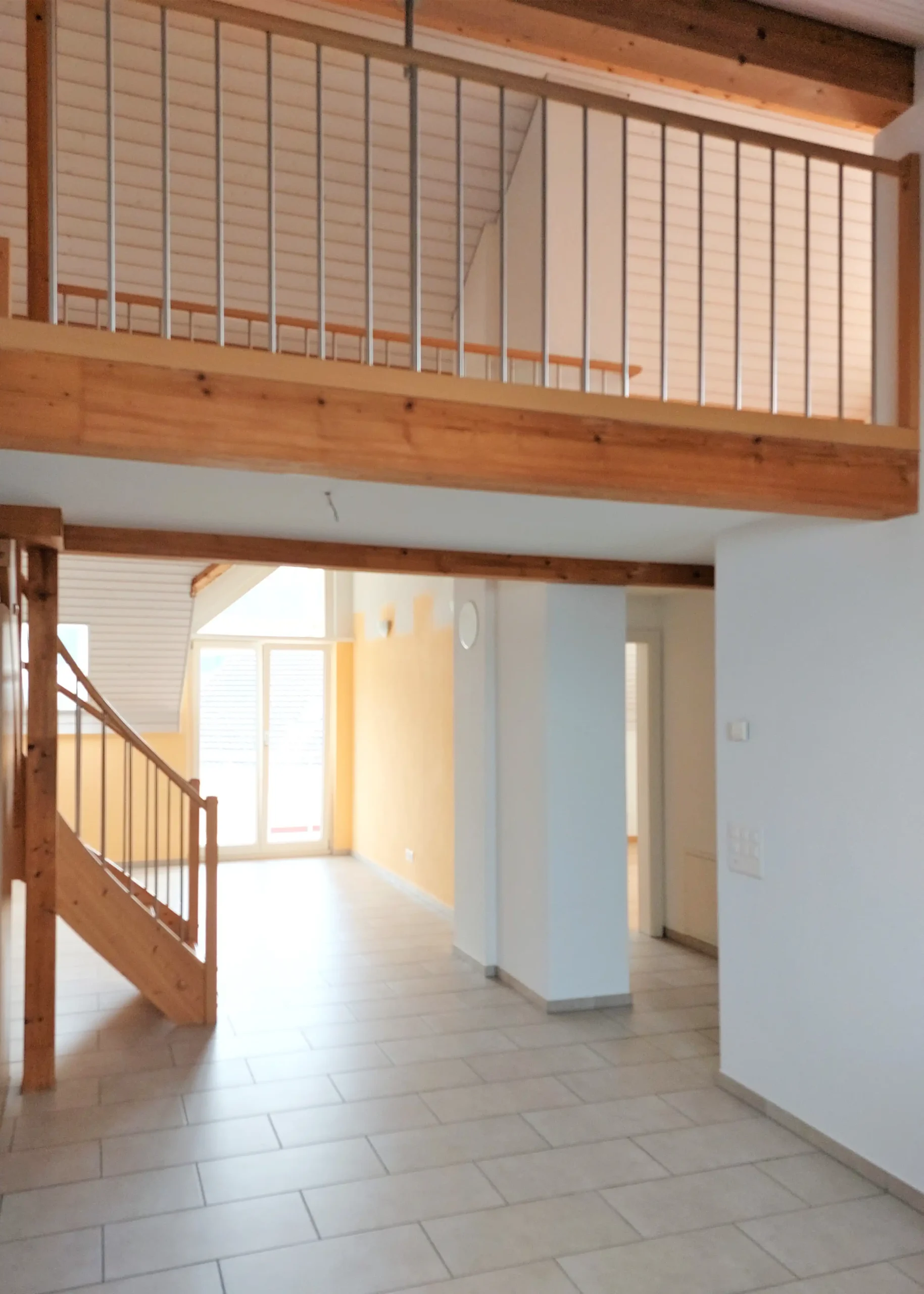 Open indoor space with tiled floor, exposed wooden beams, and a metal railing balcony along the upper level; a wooden staircase is visible on the left.