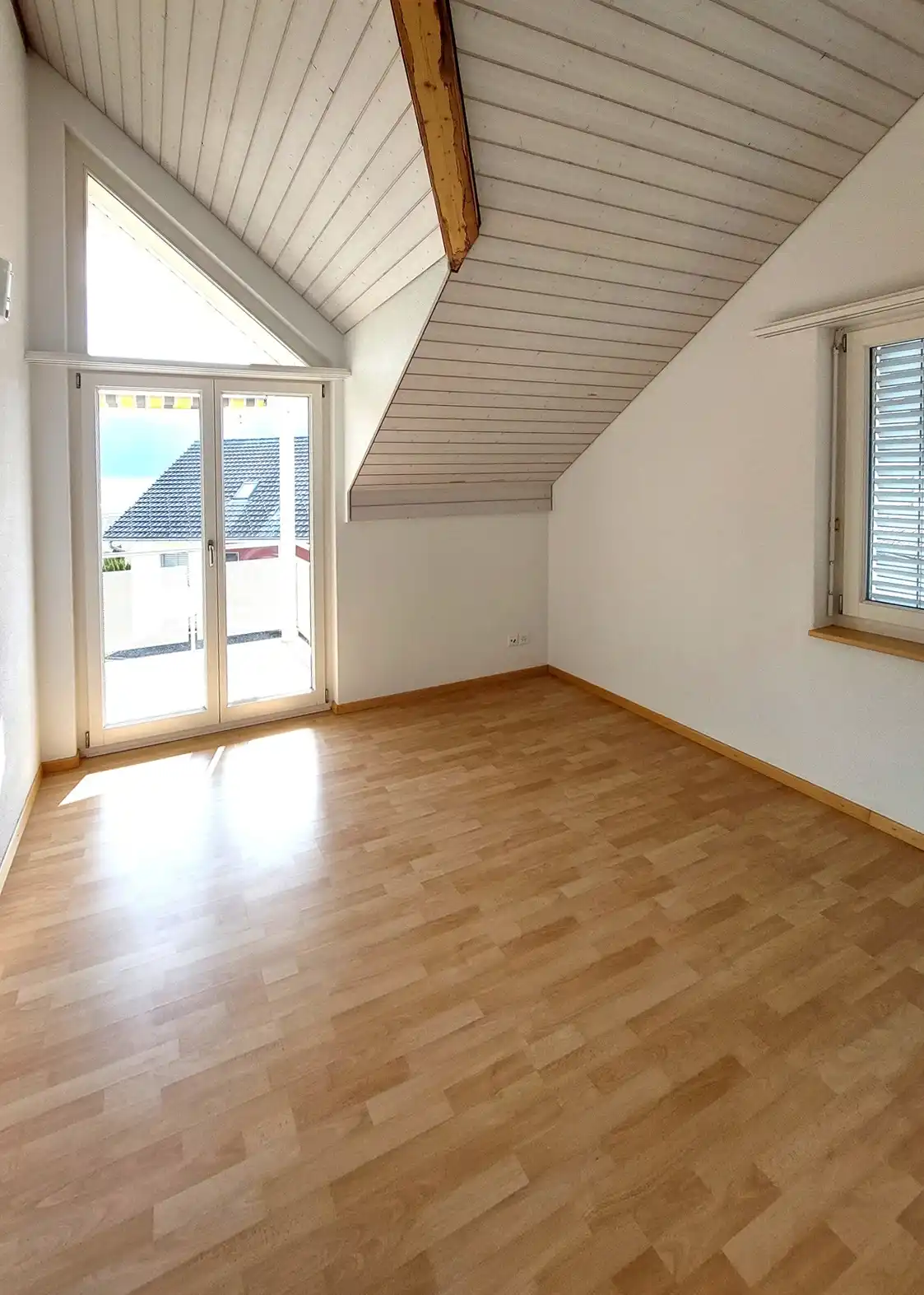 Bright empty attic room with a sloped white paneled ceiling, exposed wooden beam, and light wood flooring; French doors open to a balcony with a view of neighboring roofs.