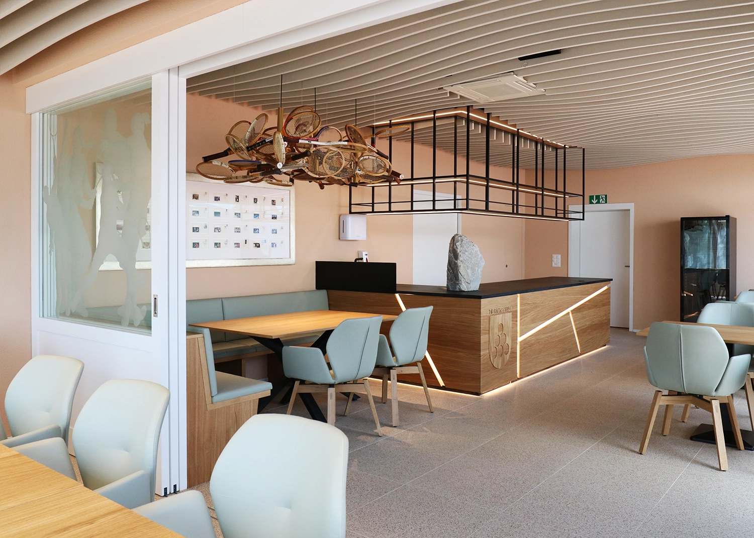 Modern cafe seating with pastel blue chairs and wooden tables; a wooden bar with a stone sculpture and a hanging rack of tennis racquets above the area.