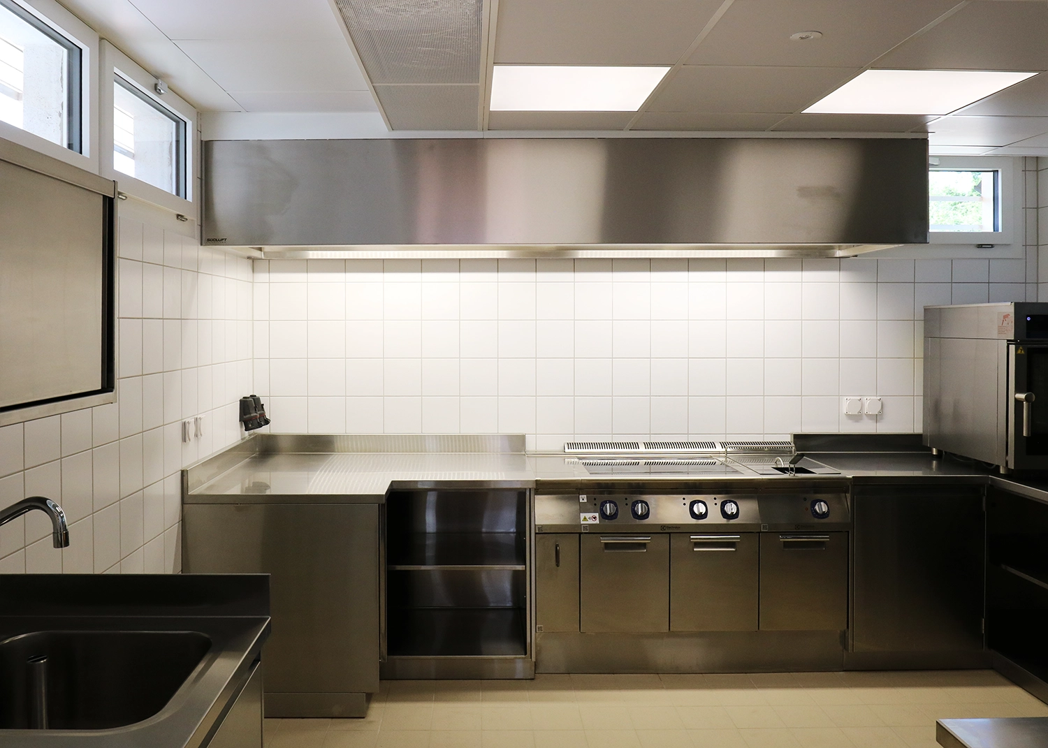 Commercial stainless steel kitchen with a large overhead exhaust hood and row of ovens and grills along a tiled wall, ready for food prep and cooking.