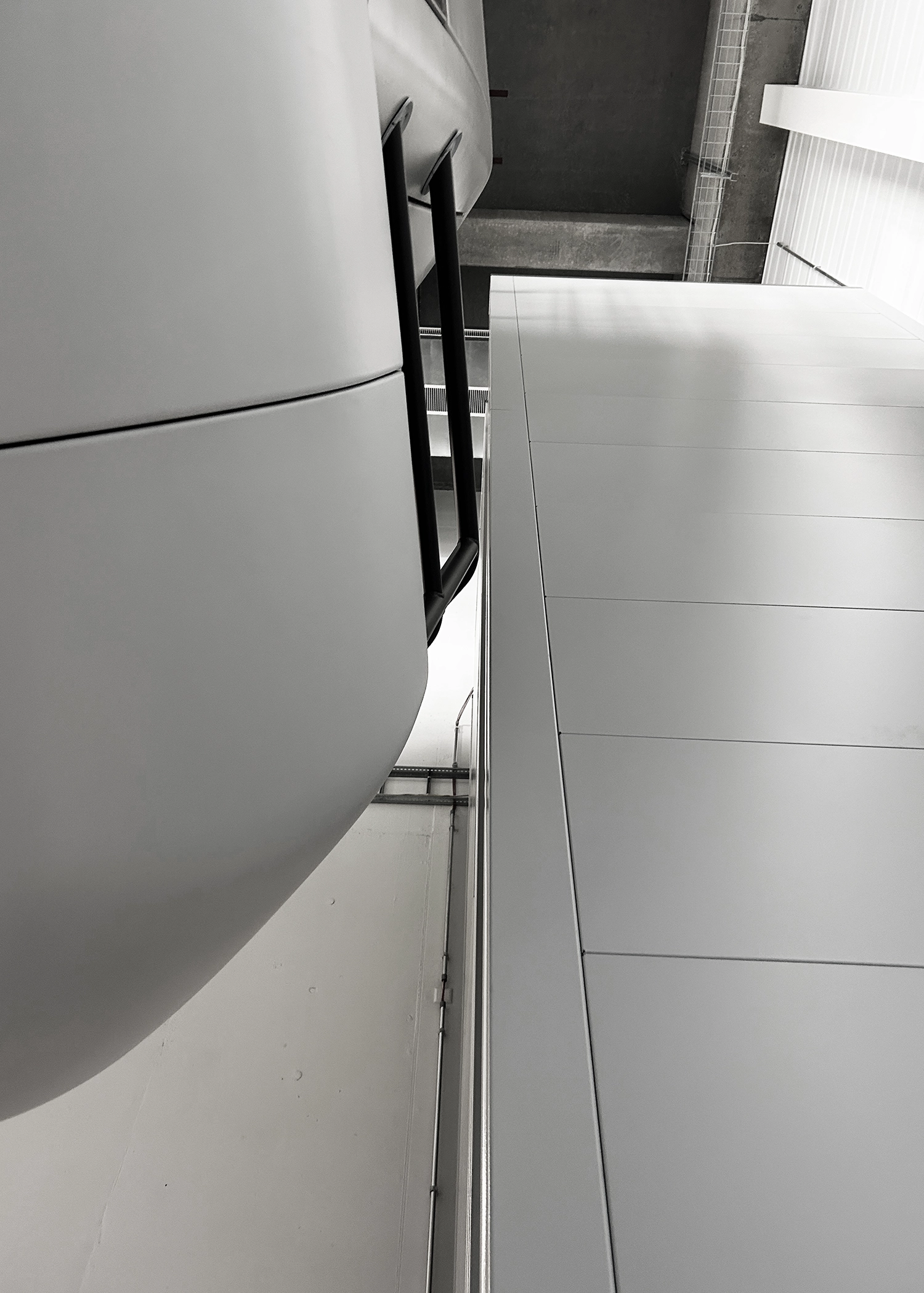 Black-and-white industrial interior shot from a low angle, showing a large curved white tank on the left and a vertical metal railing along a wall on the right.