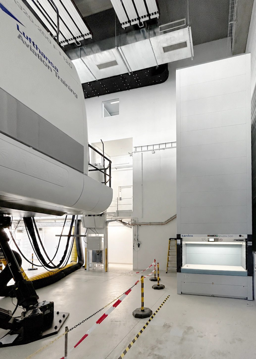 Large industrial room with a tall Kardex Shuttle 500 vertical storage unit on the right, white machinery and yellow hoses, and a taped-off restricted area.