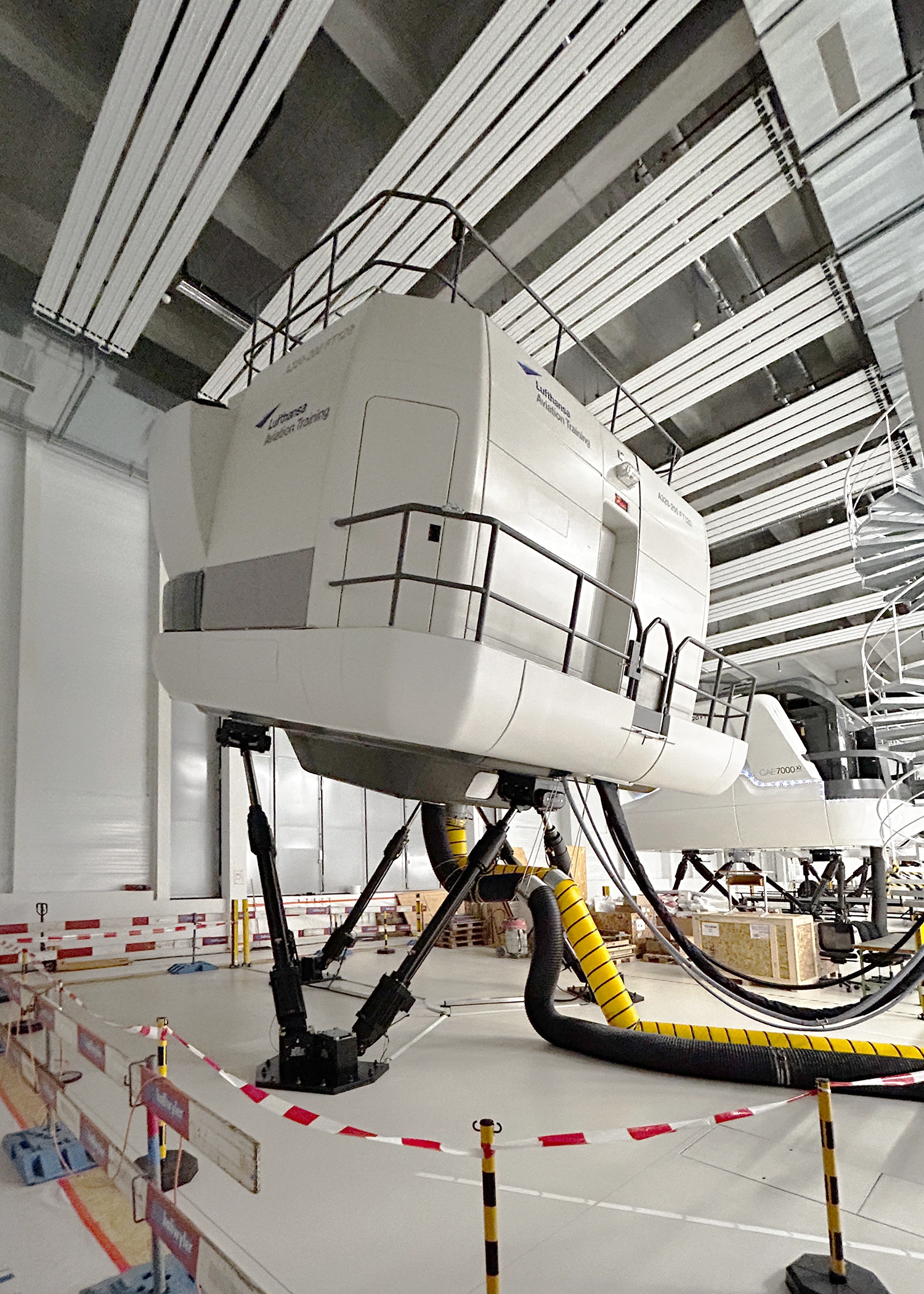 Large white aircraft flight simulator suspended in a spacious training hangar, with yellow hoses on the floor and safety barriers nearby.