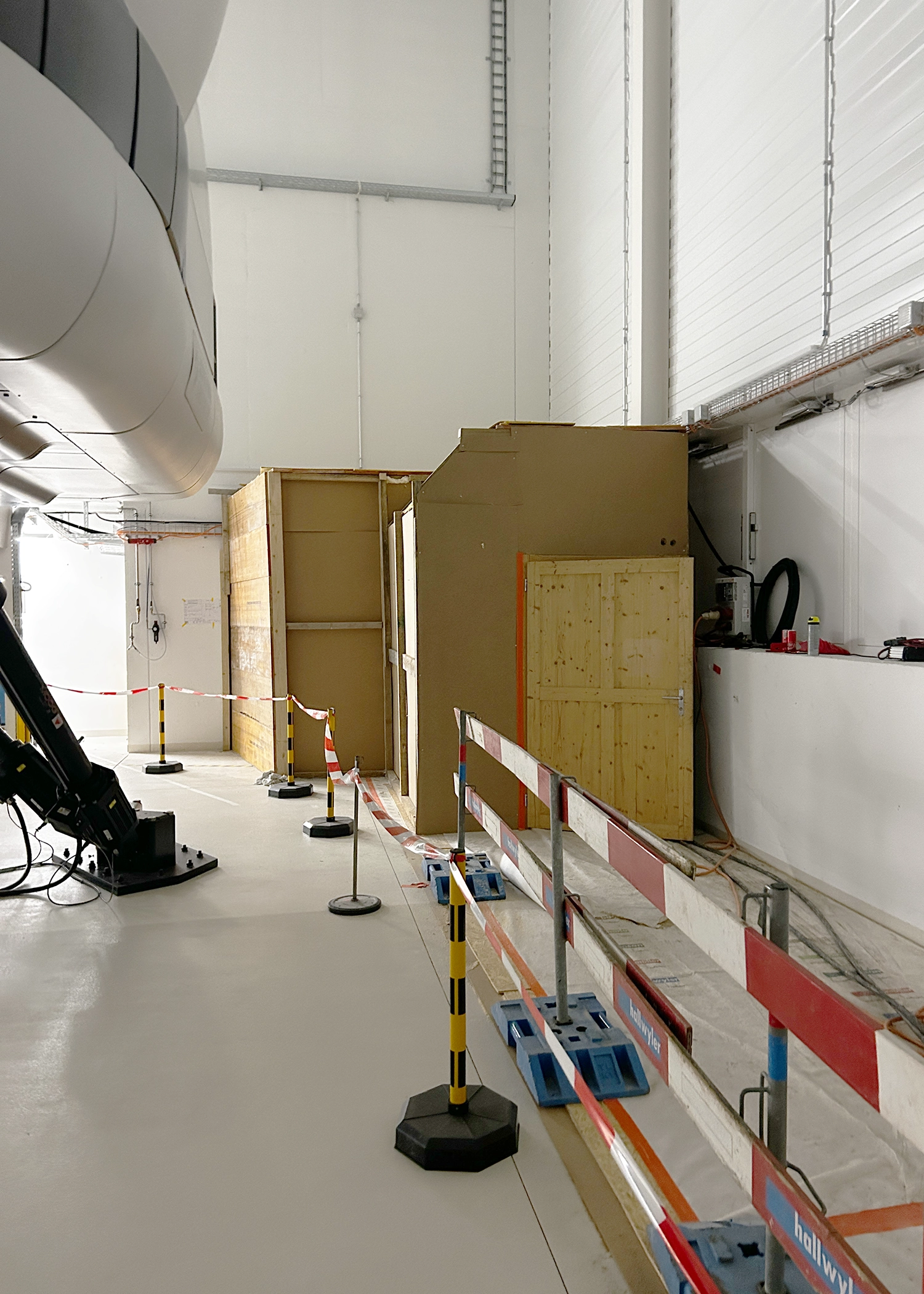 Inside a hangar with a large white aircraft fuselage section on the left, wooden crates, and red/white safety barriers along the aisle.